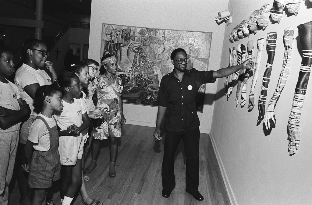 A class visiting the Studio Museum of Harlem. A teacher points at sculptures hanging on the wall as young students observe.