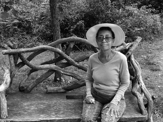 Renelda sitting on a bend in front a bush. She is wearing glasses and a wide-brimmed hat.
