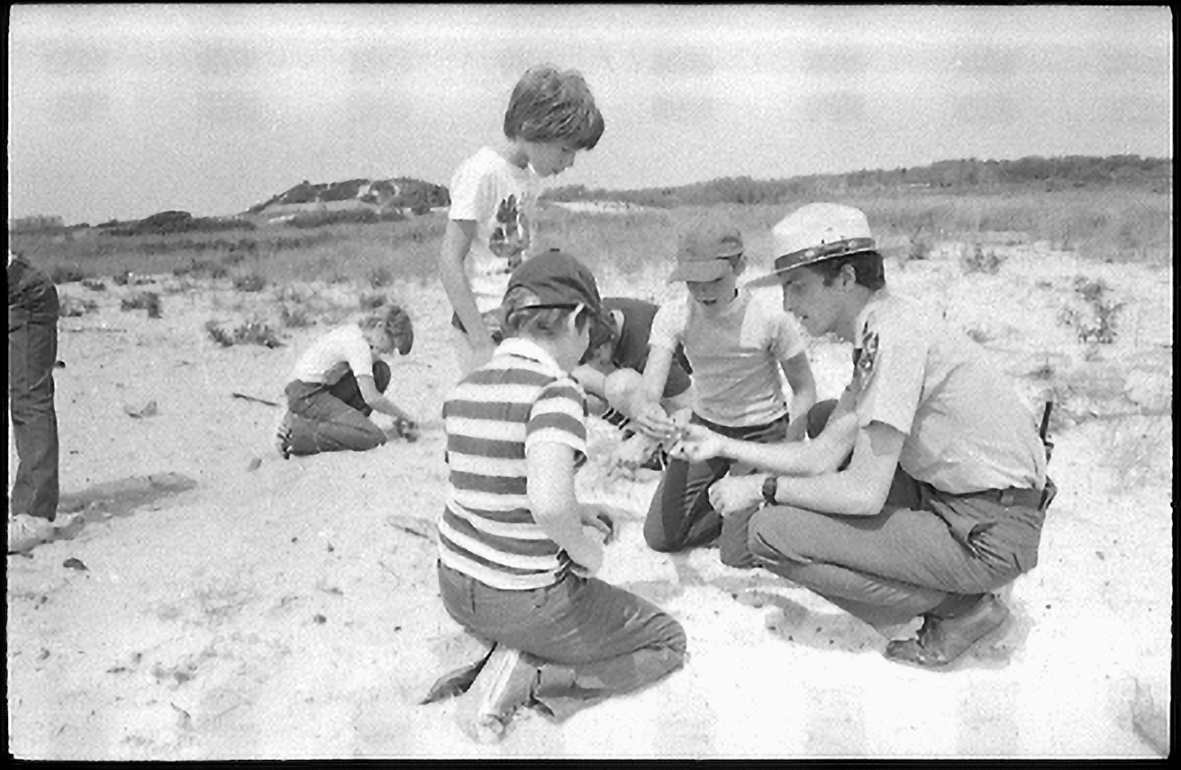 A park ranger kneeling with something in his hands. Three children look on in wonder.
