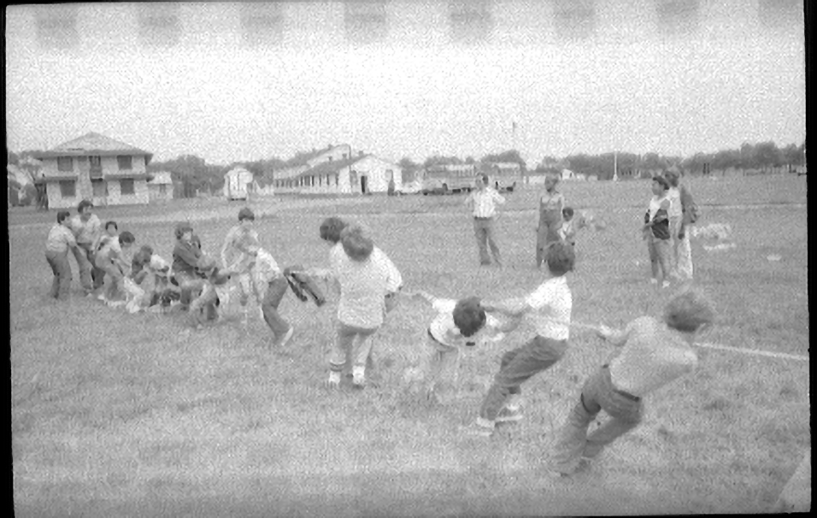 two groups of children playing tug of war in a field surrounded by houses. It's unclear who is winning. Adults look on in supervision.