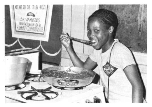 a student smiling while lifting food from a frying pan with a spoon. They are wearing a Pratt Institute t-shirt.