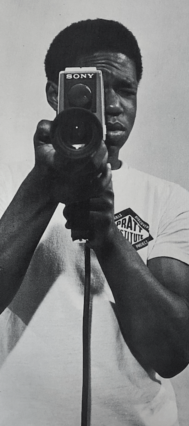 Student in a white t-shirt looking through a camera lens at the viewer