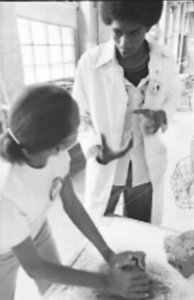 Student working with clay on a table and looking at another student gesturing