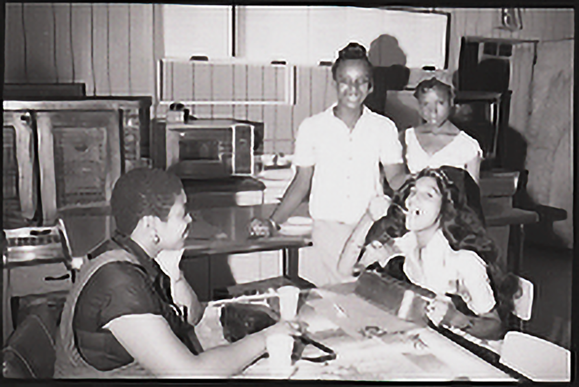 Two students seated at a table eating. Two other students smile at the camera.