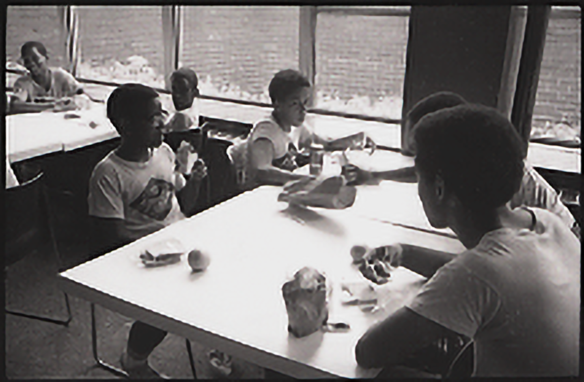 Students seated at a lunch table eating sandwiches from brown paper bags.