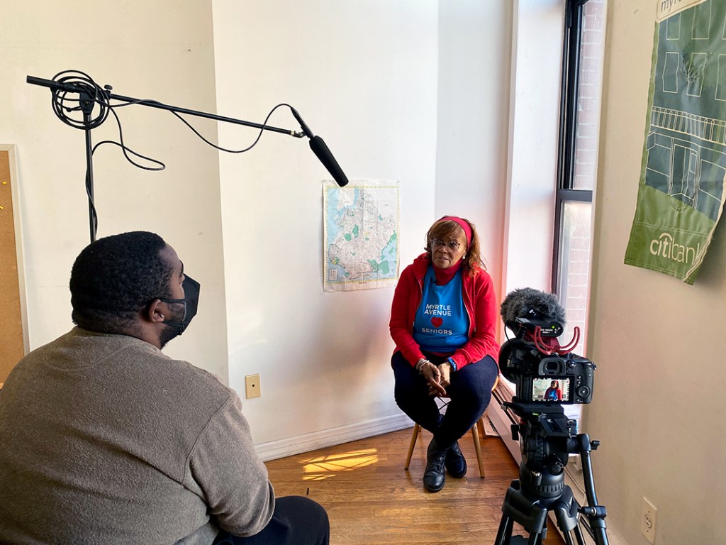 A woman in the corner of a room with an apron on that reads "Myrtle Avenue Seniors" She is being interviewed by a man with a camera and a microphone.