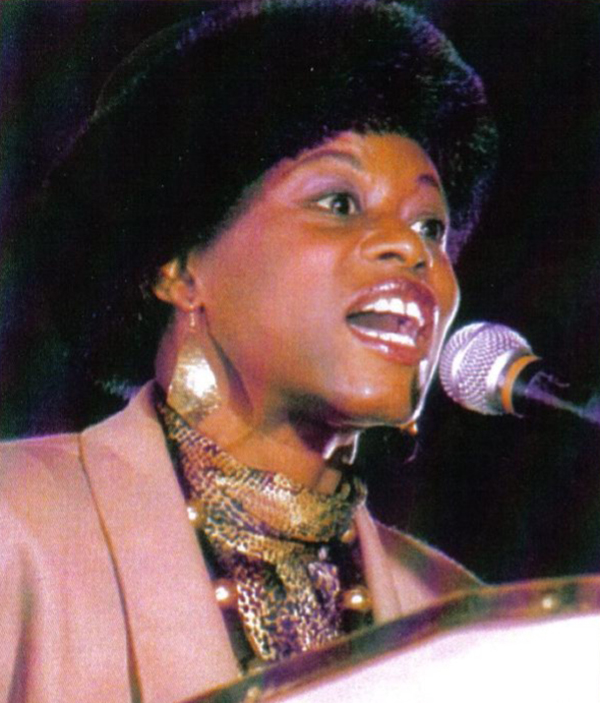 Carole Imani Parker speaking into a microphone. She is wearing a pink blazer.