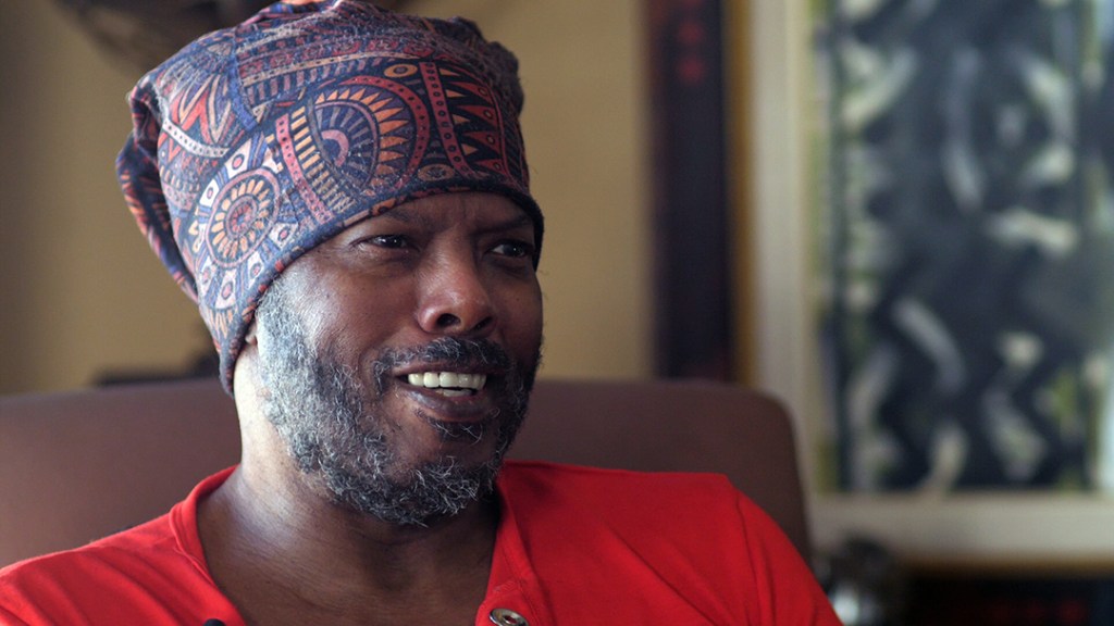 John Dew seated for an interview. He is a bearded man in a red shirt.